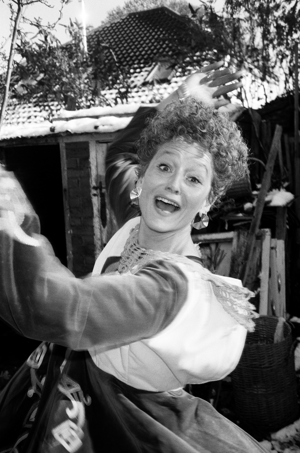 A joyful woman with curly hair and large earrings dances outside, smiling widely. She wears a dress with lace details. Behind her are a shed, plants, and a snow-dusted roof.